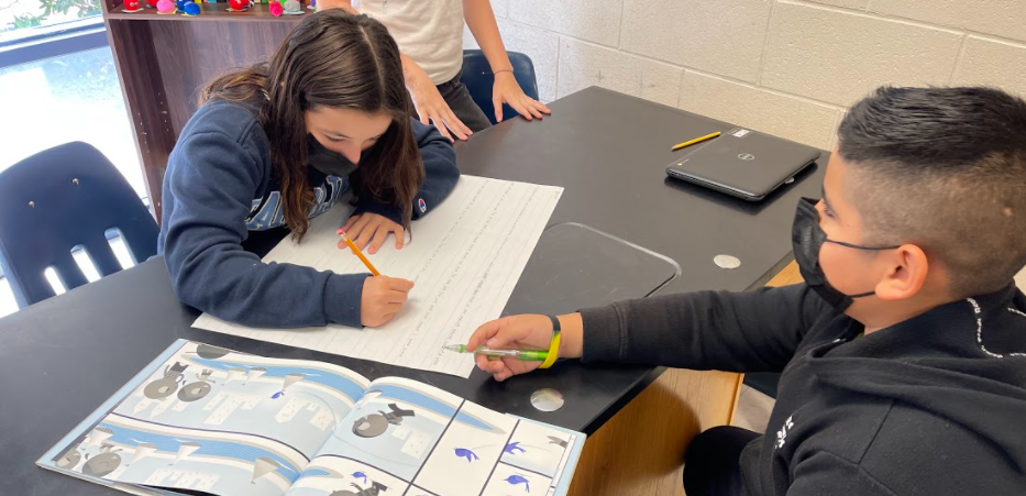 Two student are at a table.  One student is writing on a poster and another student is discussing what to write on the poster.  Another student stands in the background, looking at the poster.