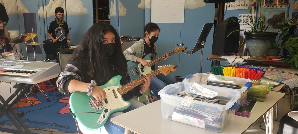 Four students (two playing drums and two playing guitar) practice afterschool at School of Rock in the Art room.