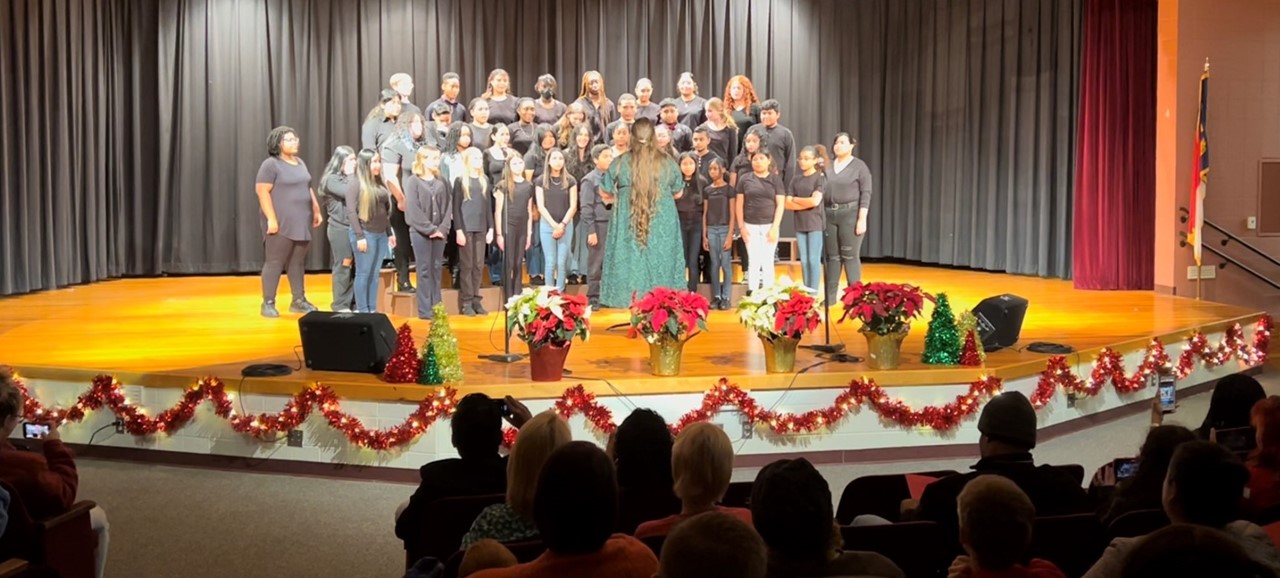 Approximately 30 students stand on three rows of risers on the center of a stage faces the audience. A teacher has her back to the crowd in front of the student  directing the chorus. The stage is decorated with red tinsel along the bottom edge facing the audience. There are four white and red poinsettias also along the front of the stage.