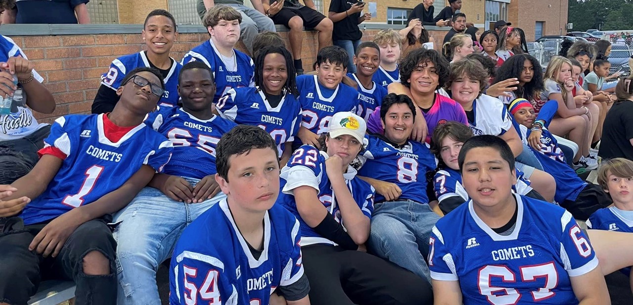 Students from football team, many in NAMS football jerseys sit in stands. Most are smiling toward the camera while some are watching the field.