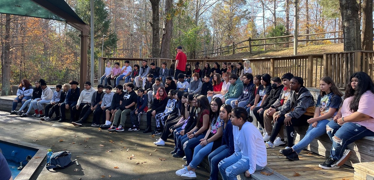 Approximately 50 students sit on three rows of risers to listen to a speaker in an outdoor area while on a fieldtrip.