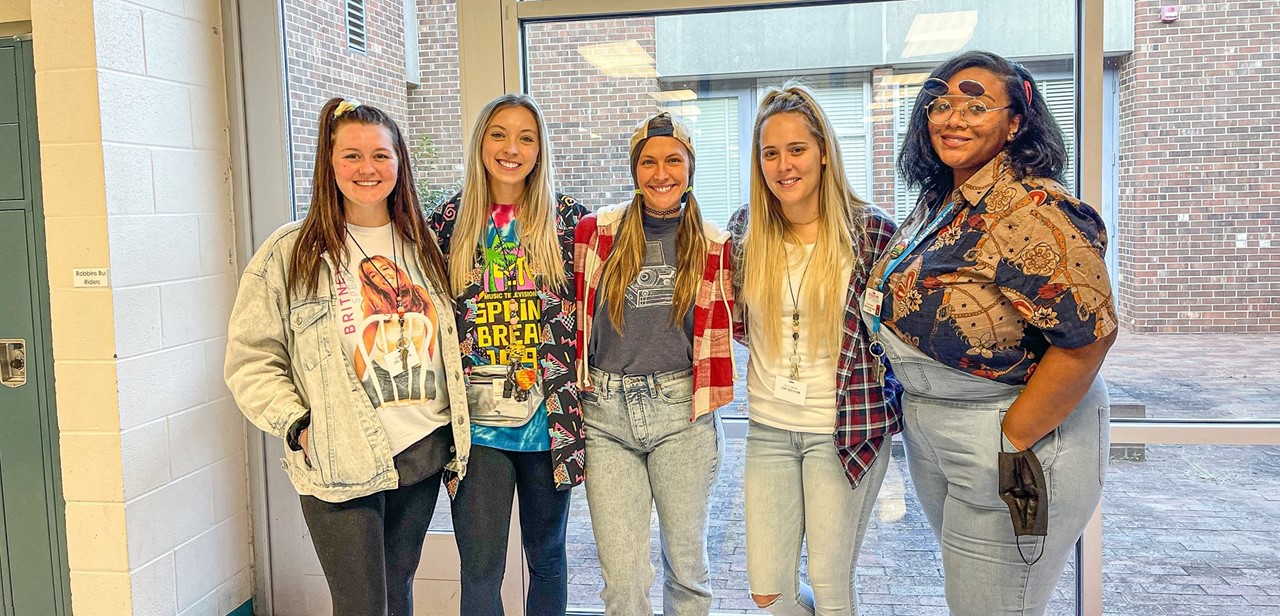 5 teachers who are dressed up for spirit week smile towards the camera while in the hallway