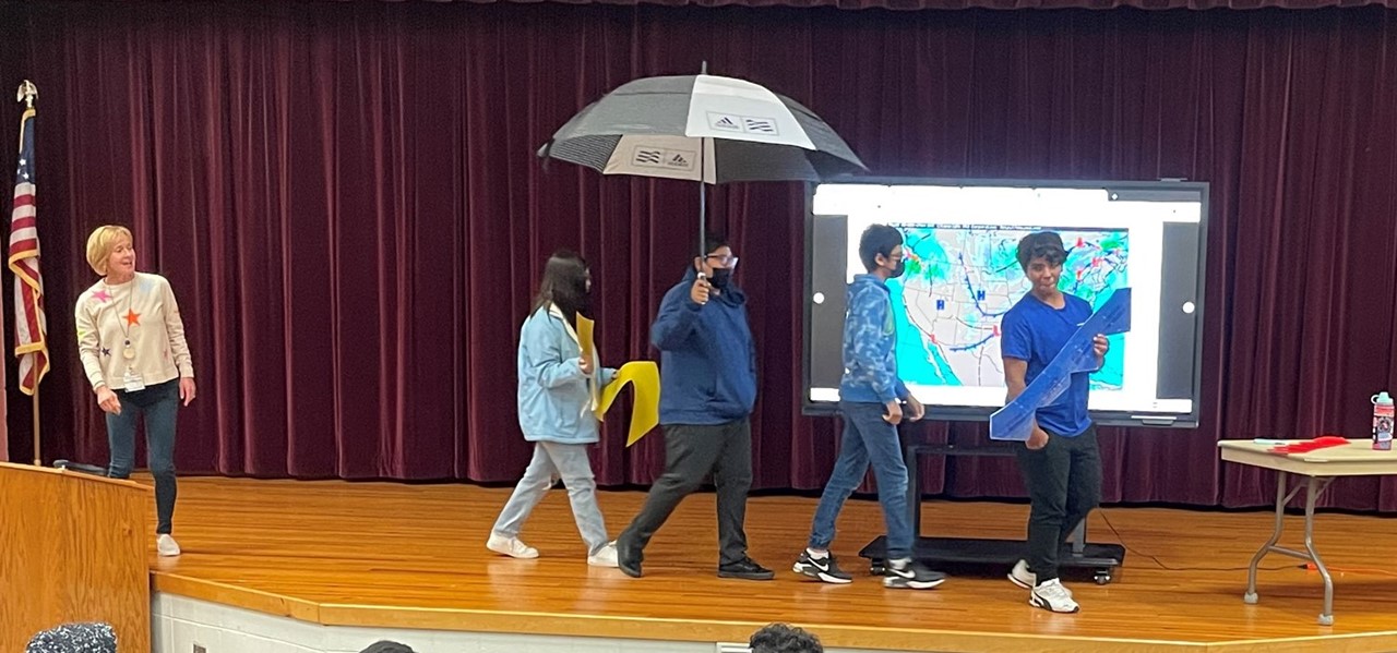 four studetns walk across the stage from left to right while carrying an umbrella and other weather items. The guest speaker is on the left and  looks towards them
