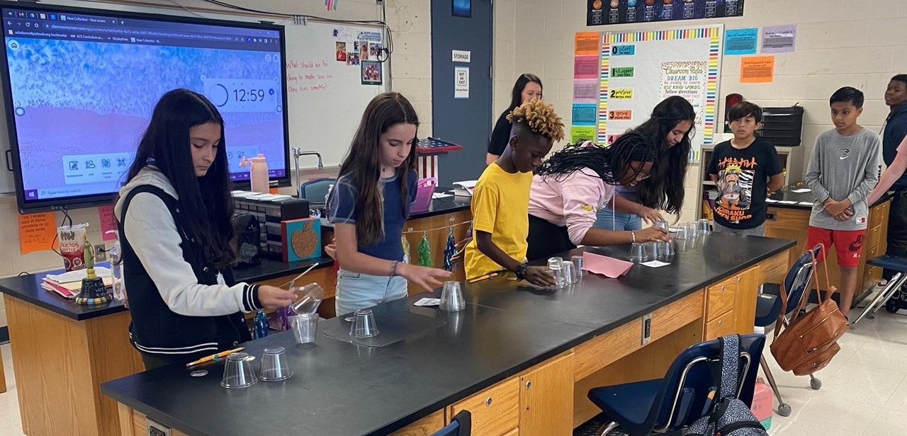 Five students stand alongside the far side of a table with clear cups and papers while working on an experiment. The teacher and three more students  observe the five students.